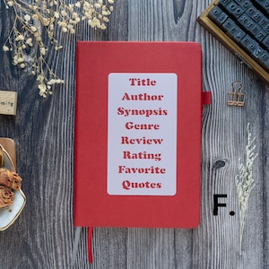Pode incluir: Um caderno vermelho com um r&oacute;tulo branco que diz "T&iacute;tulo, Autor, Sinopse, G&ecirc;nero, Revis&atilde;o, Classifica&ccedil;&atilde;o, Cita&ccedil;&otilde;es favoritas". O caderno est&aacute; sobre uma superf&iacute;cie de madeira com um pequeno bloco de madeira que diz "daydreaming" e um pequeno ramo de flores secas.