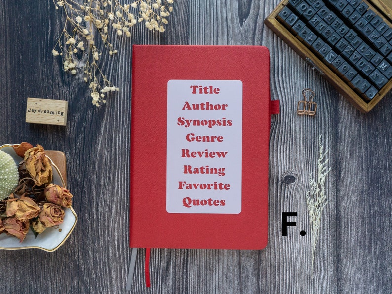 Pode incluir: Um caderno vermelho com um r&oacute;tulo branco que diz "T&iacute;tulo, Autor, Sinopse, G&ecirc;nero, Revis&atilde;o, Classifica&ccedil;&atilde;o, Cita&ccedil;&otilde;es favoritas". O caderno est&aacute; sobre uma superf&iacute;cie de madeira com um pequeno bloco de madeira que diz "daydreaming" e um pequeno ramo de flores secas.
