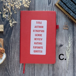 Pode incluir: Um di&aacute;rio de capa dura vermelho com um adesivo branco na frente. O adesivo tem as palavras "T&Iacute;TULO", "AUTOR", "SINOPSE", "G&Ecirc;NERO", "REVIS&Atilde;O", "CLASSIFICA&Ccedil;&Atilde;O", "FAVORITO", e "CITA&Ccedil;&Otilde;ES" impressas nele.