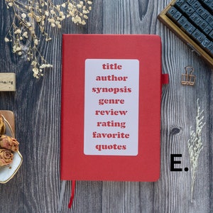 Pode incluir: Um caderno vermelho com um adesivo branco que diz "t&iacute;tulo, autor, sinopse, g&ecirc;nero, revis&atilde;o, classifica&ccedil;&atilde;o, favorito, cita&ccedil;&otilde;es". O caderno est&aacute; sobre uma superf&iacute;cie de madeira com um pequeno carimbo de madeira que diz "daydreaming" e um carimbo de metal que diz "E."