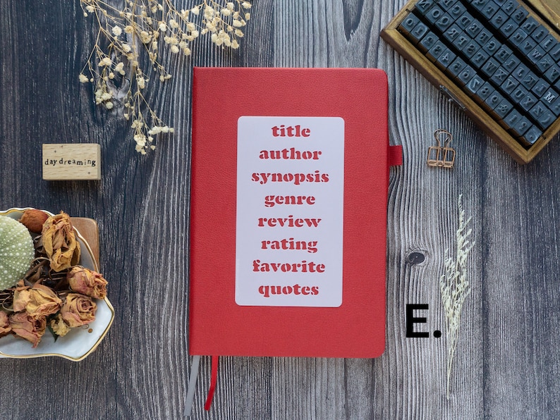 Pode incluir: Um caderno vermelho com um adesivo branco que diz "t&iacute;tulo, autor, sinopse, g&ecirc;nero, revis&atilde;o, classifica&ccedil;&atilde;o, favorito, cita&ccedil;&otilde;es". O caderno est&aacute; sobre uma superf&iacute;cie de madeira com um pequeno carimbo de madeira que diz "daydreaming" e um carimbo de metal que diz "E."