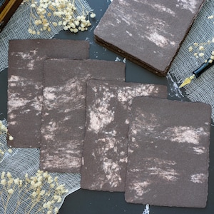 May include: Five sheets of dark brown paper with a marbled pattern. The paper has a rough texture and is perfect for writing or drawing.