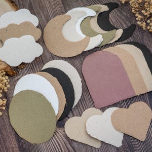 May include: Assortment of handmade paper shapes in various colors and designs, including clouds, moons, circles, arches, and hearts. The shapes are in neutral tones of white, beige, brown, black, and olive green, arranged on a wooden surface.