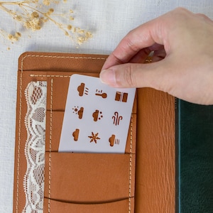 May include: A tan leather planner cover with white stitching and a white lace trim detail. A rectangular white stencil featuring nine weather-related icons, including clouds, rain, sun, and a snowflake, is tucked into a pocket on the front cover.