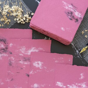 May include: A stack of pink handmade paper sheets with a speckled, marbled pattern. The paper has a rough, textured surface and is ideal for writing or crafting.