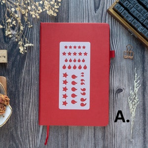 Pode incluir: Um caderno vermelho com um est&ecirc;ncil branco de estrelas, chamas e folhas. O caderno est&aacute; fechado e tem um marcador de metal prateado.