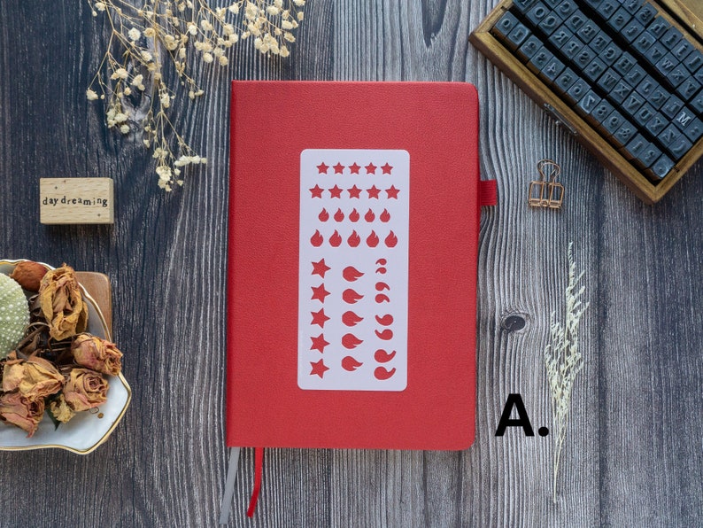 Pode incluir: Um caderno vermelho com um est&ecirc;ncil branco de estrelas, chamas e folhas. O caderno est&aacute; fechado e tem um marcador de metal prateado.