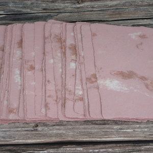 May include: A stack of handmade paper sheets in a pale pink color with brown and white marbling. The paper has a rough, textured surface and uneven edges.