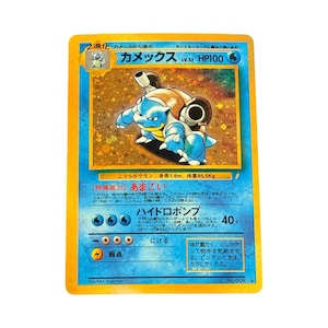 May include: A Japanese Blastoise trading card featuring the blue turtle-like creature with cannons on its back. The card has a yellow border, a blue background, and displays HP100, the name Kamex, and Japanese text detailing its special abilities.