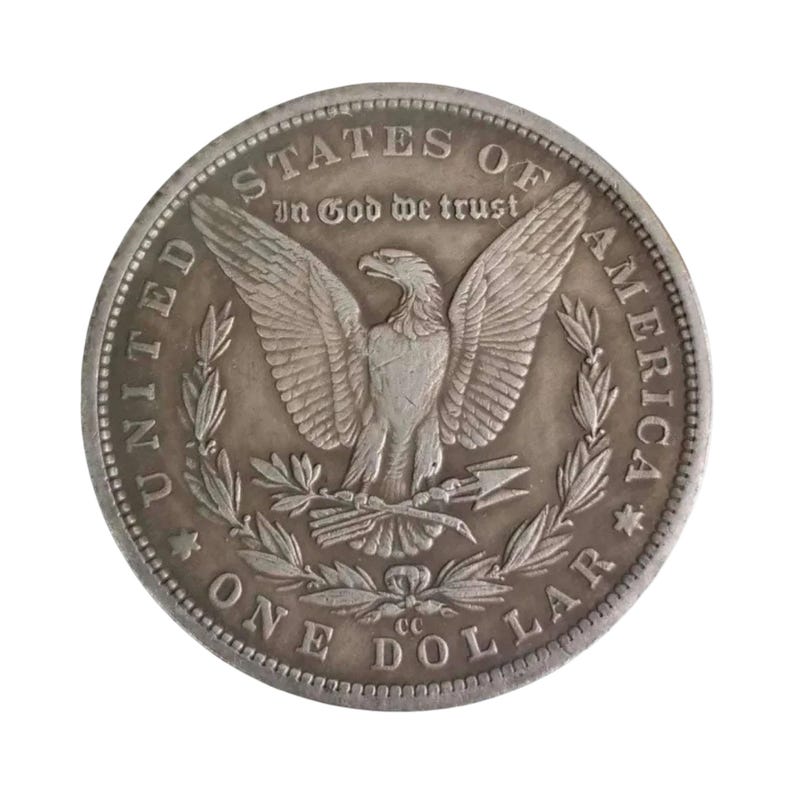Pu&ograve; includere: Una moneta da un dollaro Morgan color argento raffigurante un'aquila con le ali spiegate che stringe frecce e un ramoscello d'ulivo. Il testo recita UNITED STATES OF AMERICA, IN GOD WE TRUST, ONE DOLLAR, con il marchio CC sotto l'aquila.