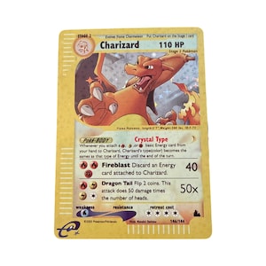 May include: A holographic Charizard trading card featuring the orange dragon-like creature with wings spread. The card displays 110 HP, the Crystal Type Poke-BODY ability, and attacks Fireblast and Dragon Tail. It is marked 146/144 and dated 2003.