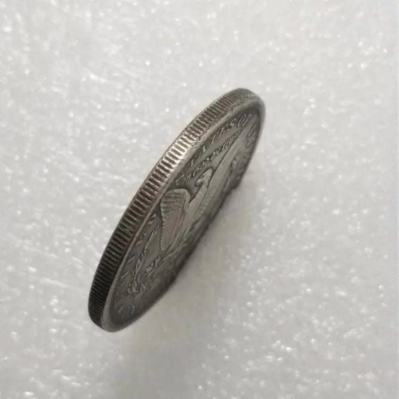 Pu&ograve; includere: Una moneta color argento vista da un'angolazione laterale, che rivela un bordo zigrinato e una vista parziale di un disegno di un'aquila sulla faccia. La moneta presenta testo metallico lungo il bordo, con forma circolare e bordo striato.