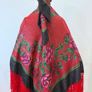 May include: A red shawl featuring a black border and a floral pattern of pink roses with green leaves. The shawl has long red fringe along the edges and is worn as a head covering with a knot at the back, displaying a vibrant, textured fabric.