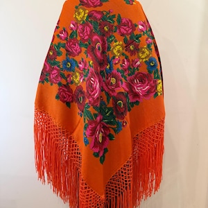 May include: A vibrant orange shawl featuring a dense floral pattern with large red, pink, and yellow roses, along with small blue flowers and green leaves. The triangular garment is finished with long, matching orange fringe along the bottom edges.