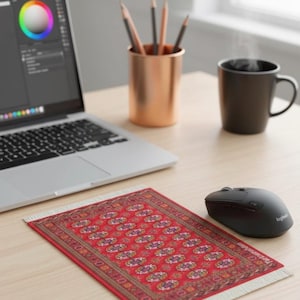 May include: A red and beige patterned rug mousepad with a white fringe sits on a wooden desk next to a black computer mouse. A laptop, pencils in a copper cup, and a black mug are also on the desk.