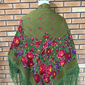 Könnte beinhalten: Ein olivgrüner Schal mit einem lebendigen Blumenmuster aus roten, rosafarbenen und violetten Rosen sowie grünen Blättern. Der Schal hat lange, geknotete Fransen an den Rändern und liegt über einer Schaufensterpuppe mit gemusterter Kopfbedeckung.