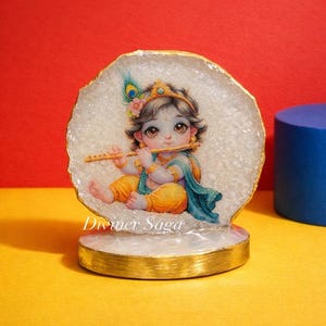 May include: A decorative figurine featuring a detailed illustration of a baby playing a flute. The figure is set against a white, textured background with a gold-colored border and base. The text "Diviner Saga" is visible.