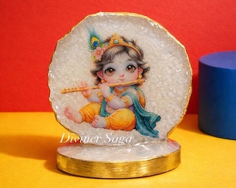 May include: A decorative figurine featuring a detailed illustration of a baby playing a flute. The figure is set against a white, textured background with a gold-colored border and base. The text "Diviner Saga" is visible.