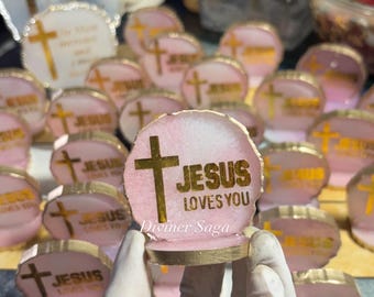 May include: Pink and gold resin art pieces with a cross and the words "JESUS LOVES YOU" in gold lettering. The pieces have a gold trim and are displayed on a surface. The image shows multiple pieces.