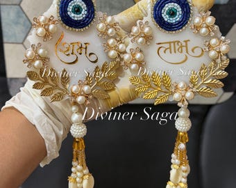 May include: A pair of decorative wall hangings featuring white circular bases with gold script text, blue and white beaded evil eye accents, clusters of white faux pearls, gold-toned metal leaf motifs, and dangling beaded tassels with white shells.