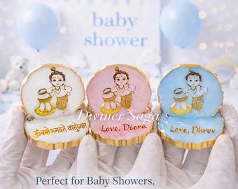 May include: Three decorative baby shower favors. Each favor features a cartoon illustration of a child with a pot. The favors are white, pink, and blue with gold trim. The text "Diviner Saga" and names are printed on the favors.