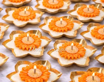 Puede incluir: Una colección de velas encendidas con forma de flor naranja en recipientes con bordes dorados en forma de pétalo. Las velas tienen una base blanca y están dispuestas sobre una superficie clara. El texto "Diviner Saga" es visible.