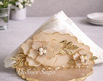 May include: A decorative napkin holder with a floral design. The holder is beige with gold accents, featuring two large flowers with pearl and rhinestone centers. The base has gold leaf details, and the text "Diviner Saga" is visible.