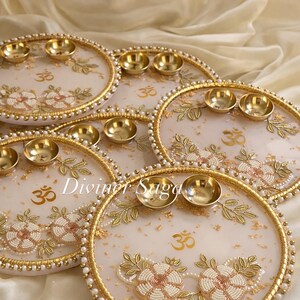 May include: Several decorative white and gold plates with floral and leaf designs, gold bowls, and the Om symbol. The plates have gold beaded edges and are arranged on a cream-colored surface. The text "Diviner Saga" is visible.