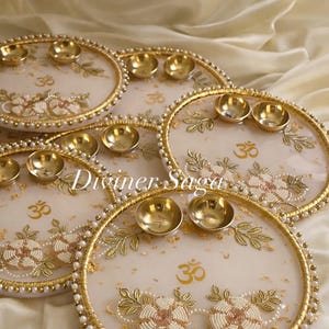 May include: Several decorative, round trays with gold rims, pearl accents, and floral designs. Each tray features small, gold bowls and the symbol "Om". The trays have a cream-colored background and the text "Diviner Saga".