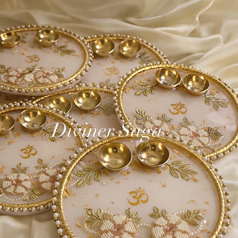 May include: Several decorative, round trays with gold rims, pearl accents, and floral designs. Each tray features small, gold bowls and the symbol "Om". The trays have a cream-colored background and the text "Diviner Saga".