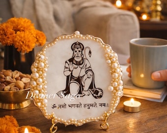 May include: A decorative, round, white and gold resin piece featuring a black illustration of a deity, surrounded by faux pearls. The piece is displayed on a gold stand. Text on the piece reads "Diviner Saga" and "ॐ नमो भगवते हनुमते नमः।".
