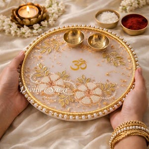 May include: A decorative white and gold tray with floral and leaf designs, featuring the Om symbol. The tray has two small gold bowls and is adorned with pearls. The tray is held by a person wearing gold bracelets.