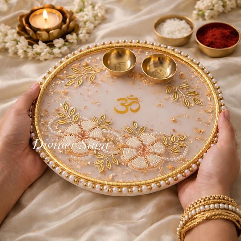 May include: A decorative white and gold tray with floral and leaf designs, featuring the Om symbol. The tray has two small gold bowls and is adorned with pearls. The tray is held by a person wearing gold bracelets.