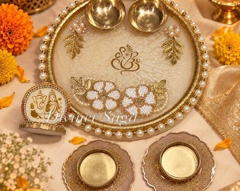 May include: A round decorative tray with a cream-colored resin base, gold-toned trim, and pearl accents. It features a central Ganesha motif, two small gold bowls, and two white beaded floral appliqués. Includes two matching coasters and a small sign.