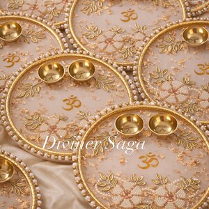 May include: Ornate, gold-colored decorative plates with floral and leaf designs, small brass bowls, and the Om symbol. The plates have a pearl-like border and the text "Diviner Saga".
