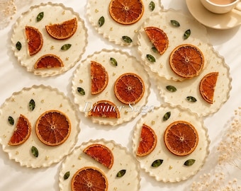 May include: A collection of decorative resin coasters with dried orange slices, green leaves, and gold flecks. The coasters have a scalloped edge and are arranged on a light-colored surface. A cup of coffee is visible in the background. The text "Diviner Saga" is visible.