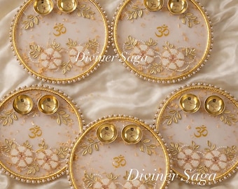 May include: Five decorative round plates with gold rims, adorned with white floral beadwork, gold accents, and small gold bowls. The plates feature the text "Diviner Saga" and the Om symbol.
