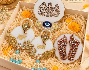 May include: A gift box containing four white resin decorative pieces. One features a line drawing of deities, one has a blue evil eye, two are circular with gold Ganesha figures and peacock accents with blue beads, and one shows red feet with clear stones.