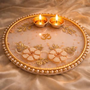May include: A decorative round tray with two lit candles. The tray has a gold rim with pearl accents and is adorned with gold floral designs and the Om symbol. The tray's surface is a creamy white color.