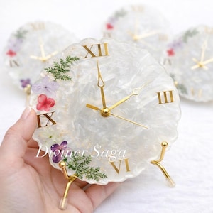 Peut inclure: Une horloge en résine blanche avec des chiffres romains et des aiguilles dorées, ornée de fleurs et de verdure séchées. L'horloge est soutenue par des pieds dorés et porte le texte "Diviner Saga".