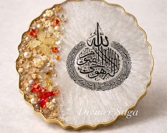 May include: Round white resin coaster with gold trim and an ornate Arabic calligraphy design. The coaster is embellished with gold, pearl, and red decorative elements. The text "Divomer Saga" is visible.
