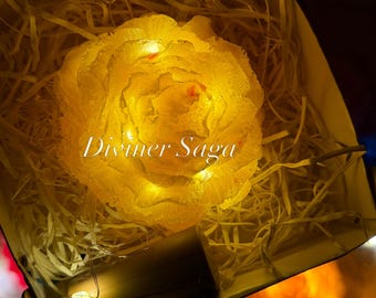 May include: A yellow rose-shaped light fixture with integrated LED string lights, nestled in a box filled with shredded paper filler. The text Diviner Saga is printed across the center of the flower. A small battery pack is visible at the bottom.