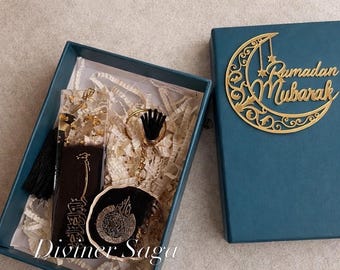 May include: A teal gift box with a gold crescent moon and "Ramadan Mubarak" text. Inside, a bookmark, keychain, and a resin pendant with gold accents are visible. The bookmark and pendant have Arabic calligraphy. The box is lined with white paper.