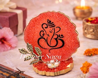 May include: A red resin coaster with a gold edge, featuring a black illustration of Ganesha. A gold leaf and pearl embellishment is attached to the coaster. The text "Dinner Saga" is visible on the coaster.
