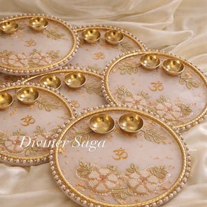 May include: Several decorative, round trays with gold rims, small gold bowls, and floral designs. The trays have a white background with gold accents, pearls, and the text "Diviner Saga" and the Om symbol.