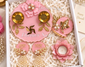 May include: A gift box containing pink resin decor items including a circular tray with two gold diya holders, a swastika symbol, and a floral accent. Includes a Ganesha figurine on a pink base, two circular plaques with Hindi text, and a lotus-shaped candle holder.
