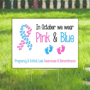 Pode incluir: Placa de jardim branca com o texto "In October we wear Pink & Blue" e "Pregnancy & Infant Loss Awareness & Remembrance". A placa tem uma fita feita de pegadas azuis e rosas.
