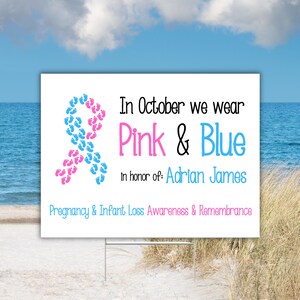 Pode incluir: Uma placa branca com o texto "In October we wear Pink & Blue" e "In honor of: Adrian James". Uma fita feita de pegadas rosa e azuis est&aacute; &agrave; esquerda. A placa est&aacute; em uma praia.