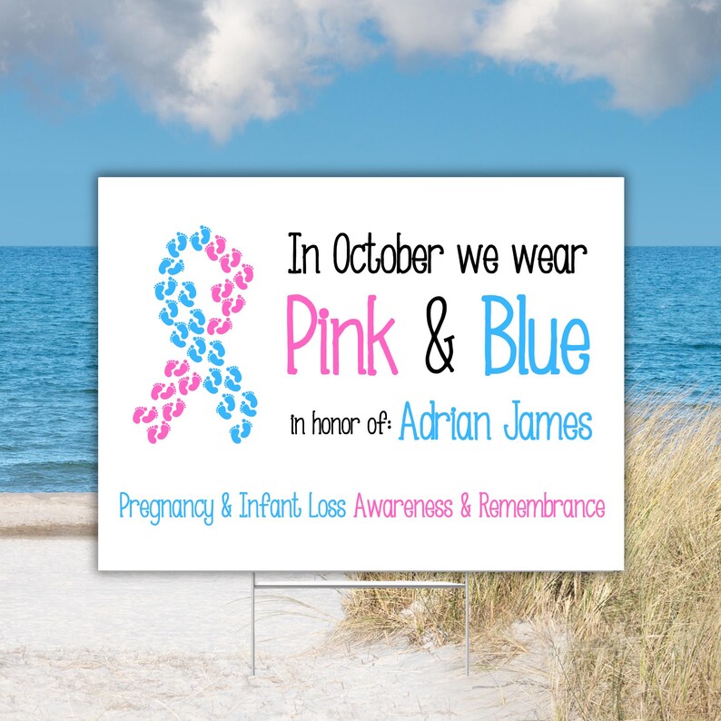 Pode incluir: Uma placa branca com o texto "In October we wear Pink & Blue" e "In honor of: Adrian James". Uma fita feita de pegadas rosa e azuis est&aacute; &agrave; esquerda. A placa est&aacute; em uma praia.