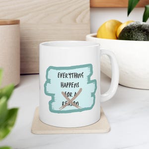 Könnte beinhalten: Weiße Keramiktasse mit dem Text "Everything Happens For A Reason" in schwarzer Schrift auf hellblauem Hintergrund. Die Tasse steht auf einem beigen Untersetzer.